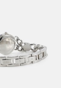Montre argentée avec un bracelet à maillons, dotée d'un boîtier rond et du logo "GUESS" gravé. Fini en métal poli et texture décorative.