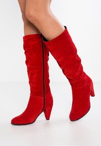 Bottes montantes en daim rouge avec un design décontracté, un bout pointu et un talon effilé. Dotées d'une fermeture éclair latérale.