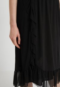 Schwarzes Kleid mit einem schulterfreiem Design auf der Seite, geschichteter Textur und einem transparenten, rüschierten Saum. Eine Hand ist sichtbar, die einen Teil des Kleides hält.