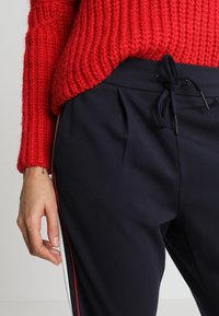 Main à côté d’un pantalon noir avec des bandes latérales rouges et blanches, portant un pull rouge en tricot avec un petit tatouage de flèche visible sur le poignet.