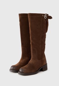 Botas altas hasta la rodilla de ante marrón con puntera redondeada, tacón bajo apilado y detalle de hebilla lateral para ajuste. Textura suave y diseño sin costuras.