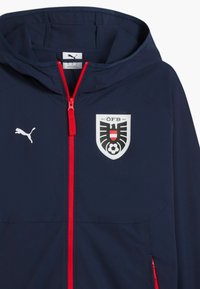 Marineblauer Kapuzenpullover mit rotem Reißverschluss, Puma-Logo auf der linken Brust und Emblem des Österreichischen Fußballbundes auf der rechten Brust.