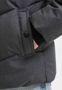 Kolfärgad pufferjacka med quiltat mönster, knappstängning på fickan och elastiska ärmslut. Tyget har ett subtilt strukturerat mönster.
