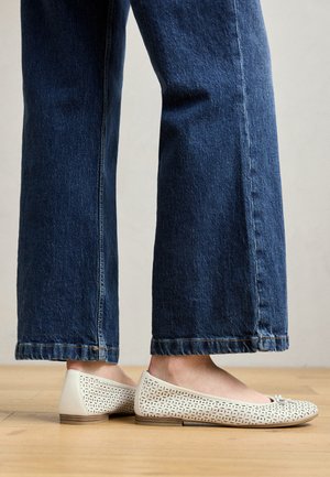 Vita perforerade ballerinas med platt sula och subtila rosett detaljer, kombinerat med uppvikt blå denim byxor med vida ben. Trägolv i bakgrunden.