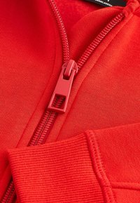 Veste zippée rouge avec un tissu lisse, des poignets côtelés et une fermeture éclair contrastante avec une tirette rectangulaire.