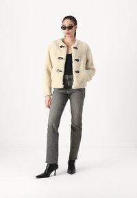 Giacca in faux fur crema con bottoni a toggles neri, abbinata a jeans grigi a vita alta e stivaletti neri. La modella indossa occhiali da sole.