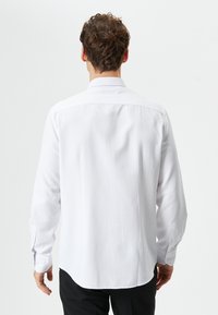 Camisa blanca de manga larga con un sutil patrón de puntos, cuello con botones y dobladillo redondeado. La tela parece suave, con textura mínima.