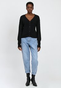 Schwarze gestrickte Strickjacke mit V-Ausschnitt und strukturierten Ärmeln, mit Knopfverschluss, kombiniert mit hellblauen, umgeschlagenen Jeans und schwarzen, hohen Stiefeln.