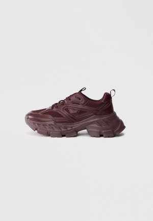 Dunkelroter klobiger Sneaker mit dicker Sohle, Mesh- und Lederpaneelen sowie Zuglaschen an Zunge und Ferse vor einfachem hellem Hintergrund.