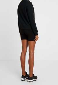 Una persona se encuentra de espaldas, vistiendo un sudadera negra, pantalones cortos ajustados negros y zapatillas negras con suelas blancas sobre un fondo blanco liso.