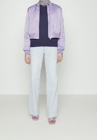 Osoba ve fialovém saténovém bomberu, tmavém námořnickém topu, světle modrých širokých džínách, fialových sandálech a zlatém řetízku.