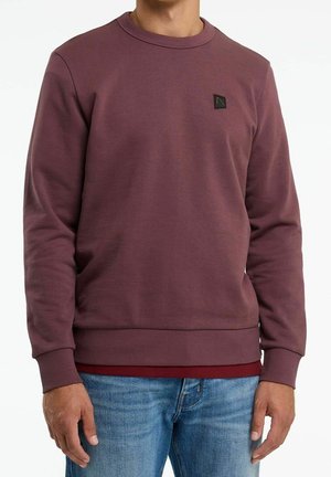Mand iført vinrød crewneck-sweatshirt over en rød skjorte, kombineret med blå jeans, vist fra hals til hofter mod hvid baggrund.