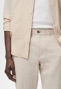 Personne à la peau foncée portant un pantalon en lin beige, une chemise à manches longues beige aux manches retroussées, et un débardeur blanc côtelé, debout avec le bras détendu.