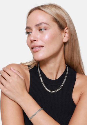 Collana e bracciale in argento su un modello, con una finitura liscia e lucida e un design classico a maglia, su sfondo di un top nero.