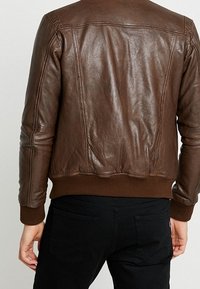 Osoba oblečená v přiléhavé hnědé kožené bundě a černých kalhotách, zobrazená zezadu, stojící proti jednoduchému světlému pozadí.