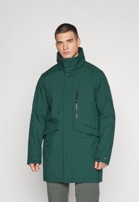 Grønn vanntett parka med høy krage, glidelåslomme på brystet, og store frontlommer. Laget av glatt, slitesterkt stoff.