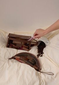 Due borsette cosmetiche marroni, una rettangolare con una parte superiore trasparente e una semicircolare. Una tazza bianca rovescia chicchi di caffè su una superficie color crema.