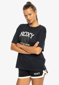 Black cotton T-shirt with white and green "ROXY 90 Active By Nature" print. Paired with black shorts featuring white accents and logo.