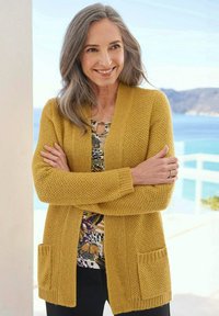 Lächelnde Frau mit grauem Haar, die einen senfgelben Strickcardigan und eine blumige Bluse trägt, steht mit verschränkten Armen an einem Balkon am Meer.