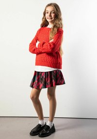 Czerwony sweter o fakturze, pod spodem biała koszula, spódnica w kratę w kolorach czerwonym, czarnym i zielonym. Czarne mokasyny z białymi skarpetkami, stojąca na jednolitym tle.