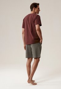 Marońska koszulka z krótkim rękawem w połączeniu z czarno-białymi szortami w kratę, wykonana z bawełny. Model stoi boso, odwrócony plecami.