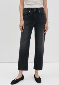 Vrouw in zwarte rechte jeans die tot de enkels zijn geknipt, met zwarte platte schoenen en een witte mouwloze top, staande tegen een effen achtergrond.