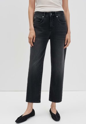 Vrouw in zwarte rechte jeans die tot de enkels zijn geknipt, met zwarte platte schoenen en een witte mouwloze top, staande tegen een effen achtergrond.