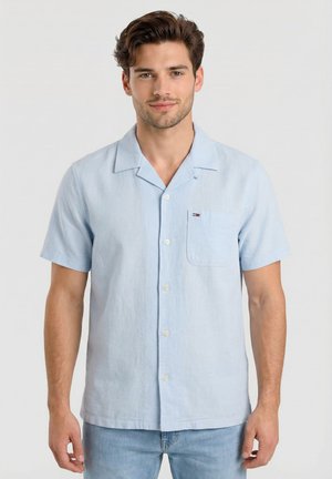 Uomo che indossa una camicia azzurra a maniche corte con colletto e taschino sul petto, abbinata a jeans azzurri, in piedi davanti a uno sfondo semplice.