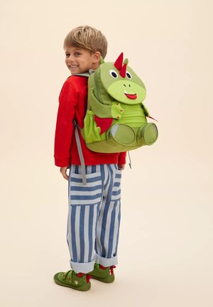 Junge im roten Sweatshirt und gestreiften Hosen lächelt, trägt einen grünen Rucksack mit Dinosaurier-Motiv, roten Hörnern und Flügeln.