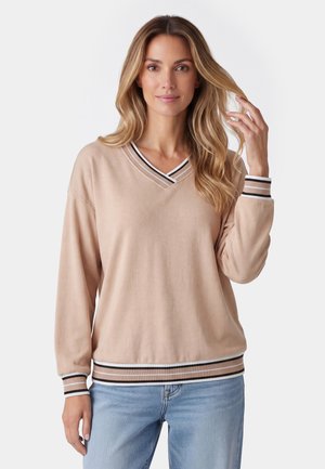 Frau mit langen blonden Haaren, die einen beigen Pullover mit V-Ausschnitt sowie gestreiften Bündchen und Saum trägt, kombiniert mit hellblauen Jeans, berührt ihr Haar.