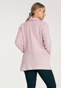 Jasnoróżowy sweter o teksturze z wykładanym kołnierzem, przednimi kieszeniami i długimi rękawami. Materiał wygląda na miękki i przytulny, idealny do noszenia na warstwy.