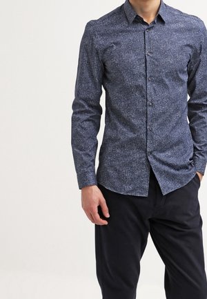 Mand iført en tætsiddende, mørkeblå skjorte med mønster og knapper, med lange ærmer og sorte bukser, stående med den ene hånd i lommen.