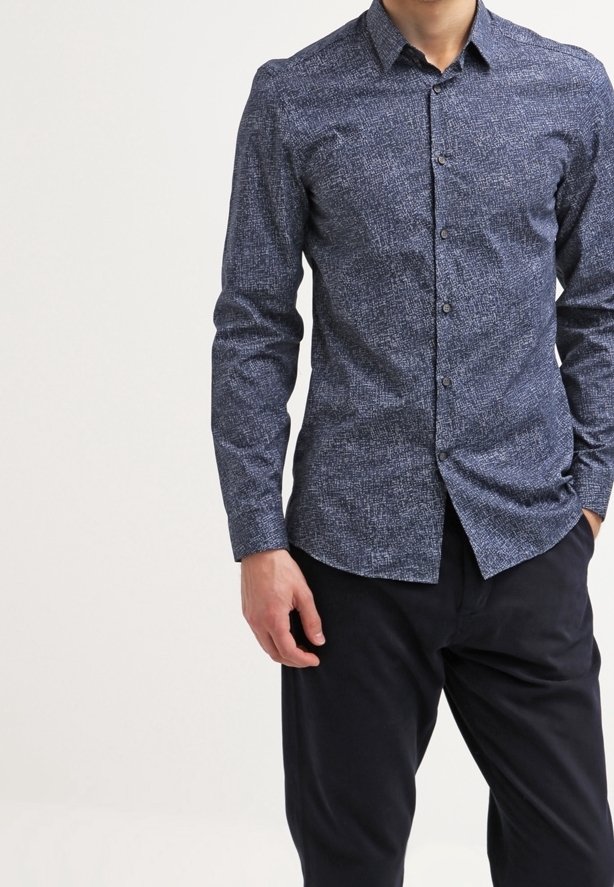 Homme portant une chemise ajustée à motifs bleu foncé à manches longues et un pantalon noir, debout avec une main dans la poche.