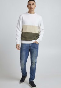 Sudadera tipo crewneck en blanco con rayas horizontales beige y verde oliva. Combinada con jeans de mezclilla azules y zapatillas negras. Textura suave.