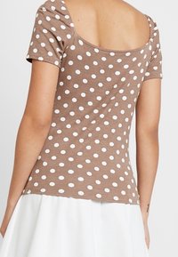 Top marrón de manga corta con lunares blancos, que presenta un diseño de escote en la espalda. Fabricado con un tejido suave y liso.