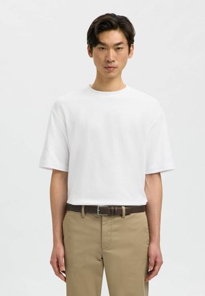 Man standing facing forward wearing a plain white short-sleeve t-shirt, beige pants, and a dark brown belt against a white background.