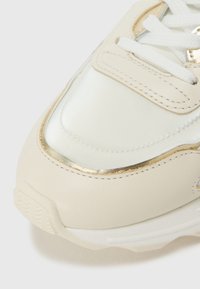 Sneaker color crema con finitura lucida, accenti metallici dorati, pelle testurizzata e suola in gomma bianca, con lacci e dettagli di cucitura.