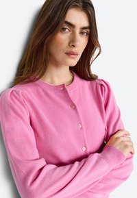 Femme aux longs cheveux bruns portant un pull rose boutonné, les bras croisés, regardant légèrement sur le côté devant un fond blanc.