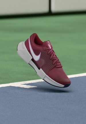 Vinrød atletiksko med hvid Nike-swoosh i luften over blå og grønne tennisbane linjer.