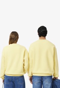 Geel crewneck sweatshirt van zacht materiaal, met ribgebreide manchetten en onderkanten, gecombineerd met blauwe denim jeans. Gezien vanuit de achterzijde.