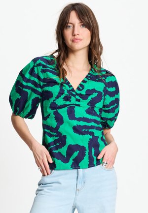 Vrouw draagt een blouse met groene en navyblauwe abstracte print, met geplooide korte mouwen en lichtblauwe jeans, handen in de zakken.