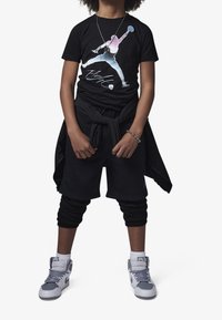 Black graphic t-shirt featuring a multicolor silhouette. Pair of black shorts with a loose fit. Grey sneakers with white accents, and a black jacket tied at the waist.