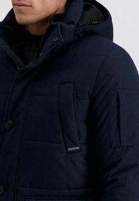 Navy blauwe gewatteerde jas met capuchon, drukknopen, twee voorzakken en gestructureerde stof. Verzien van een klein merklabel.