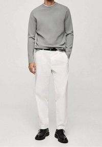 Mann in einem hellgrauen Langarmpullover, weißen Hosen, schwarzem Gürtel und schwarzen Schnürschuhen, der vor einem schlichten Hintergrund steht.