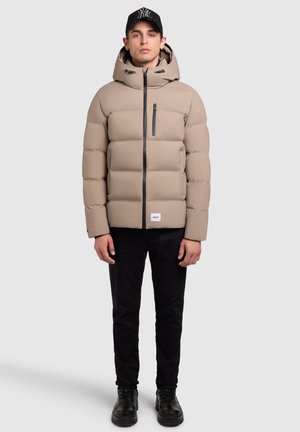 Beige pufferjack met een capuchon, zwarte ritssluiting en accenten, voorzien van een vierkant patchlogo; gedragen met zwarte broek en zwarte laarzen.