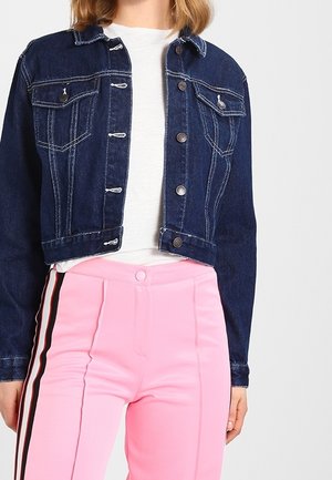 Personne portant une veste en jean bleu foncé par-dessus un haut blanc avec un pantalon rose taille haute avec des bandes latérales noires et blanches.