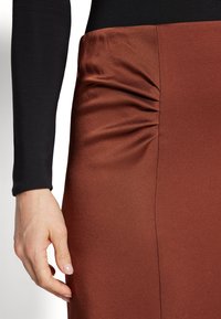Brun satengskjørt med sidedetaljer i rynker og glatt tekstur, kombinert med en svart langermet topp. En hånd berører forsiktig skjørtet.