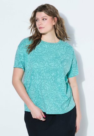 Frau mit welligem braunem Haar, die ein türkisfarbenes kurzärmliges Hemd mit Blumenmuster und einen schwarzen Rock trägt, schaut nach links vor einem weißen Hintergrund.