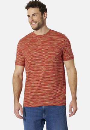 Mann lächelt, trägt ein rot-orange gestreiftes Kurzarm-T-Shirt mit Brusttasche und blaue Jeans, steht vor einfarbigem Hintergrund.