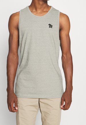 Mann in einem hellgrauen ärmellosen Tanktop mit dünnen Streifen und einem kleinen schwarzen gestickten Logo auf der Brust, kombiniert mit beigen Cargo-Shorts.